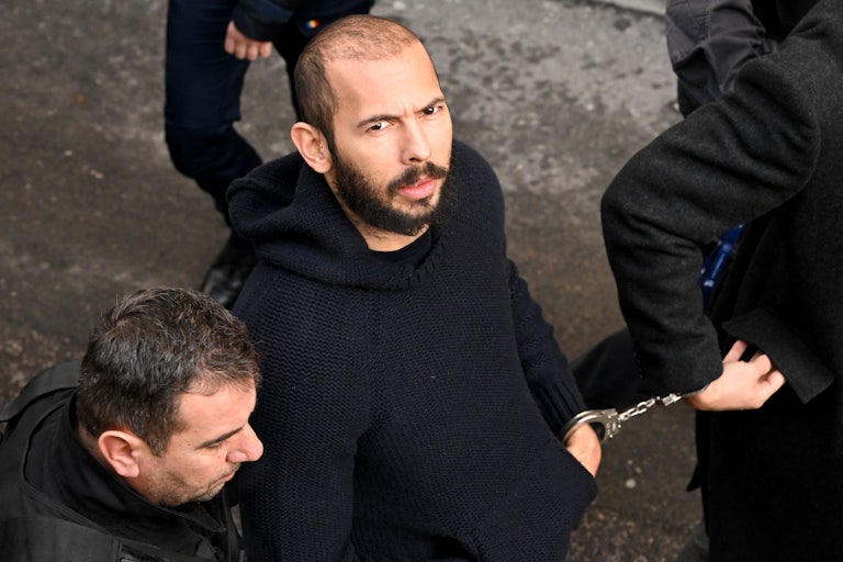 Andrew Tate, handcuffed to another man, looks up overhead directly at the camera. (He is balding.)