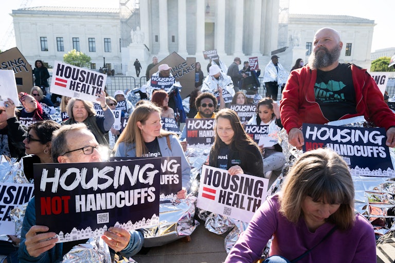At least two dozen protesters sit in front of the Supreme Court. They wear foil emergency blankets and hold signs that read "Housing = Health" and "Housing Not Handcuffs."
