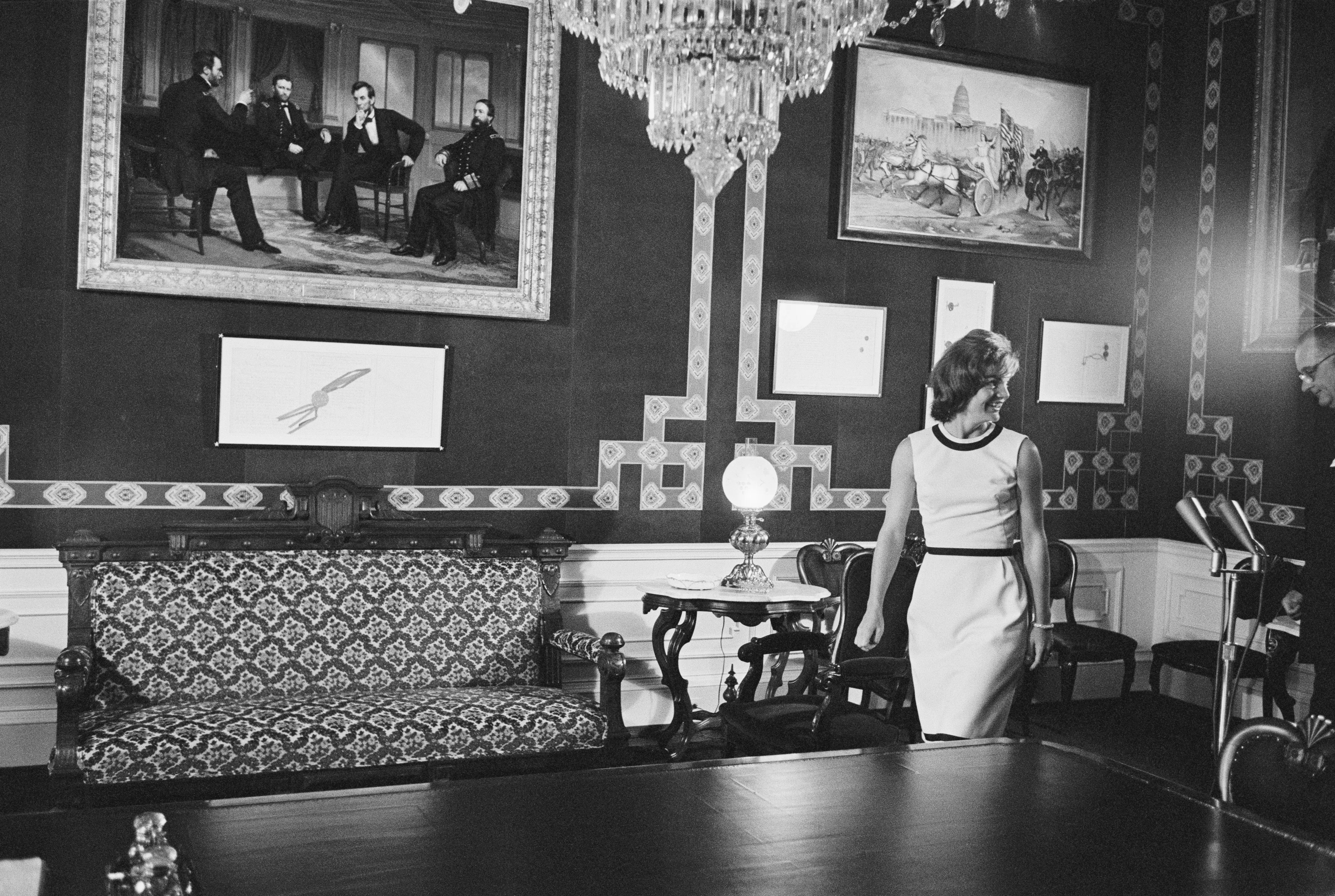 Jacqueline Kennedy in black-and-white walks into the White House Treaty Room with a picture of President Lincoln and three other people on the wall, as well as a table and sofa. A chandelier is hanging from the ceiling.