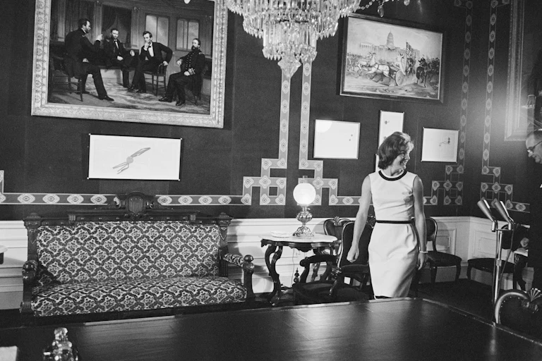 Jacqueline Kennedy in black-and-white walks into the White House Treaty Room with a picture of President Lincoln and three other people on the wall, as well as a table and sofa. A chandelier is hanging from the ceiling.