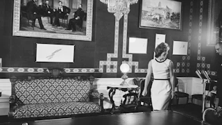 Jacqueline Kennedy in black-and-white walks into the White House Treaty Room with a picture of President Lincoln and three other people on the wall, as well as a table and sofa. A chandelier is hanging from the ceiling.
