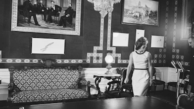 Jacqueline Kennedy in black-and-white walks into the White House Treaty Room with a picture of President Lincoln and three other people on the wall, as well as a table and sofa. A chandelier is hanging from the ceiling.
