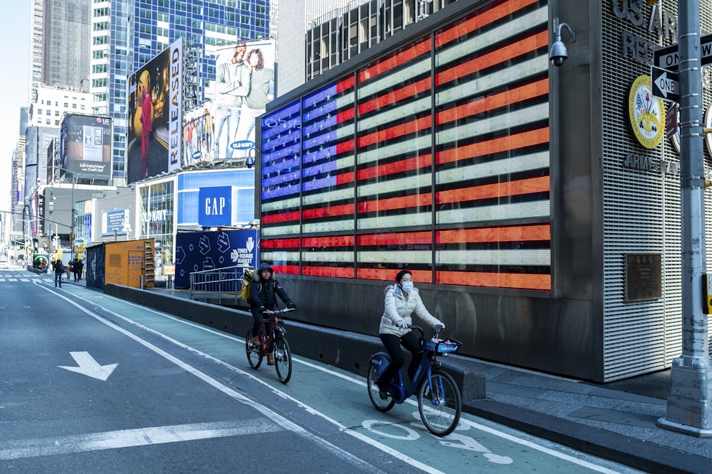 One bicyclist wearing a mask and another not wearing a mask make their way down 8th Avenue in Times Square on a Sunday afternoon with the American Flag behind it. The streets were mostly empty on the same day that New York State Governor Andrew Cuomo pleads with New York residents to take the stay-at-home orders seriously regarding the Coronavirus pandemic.