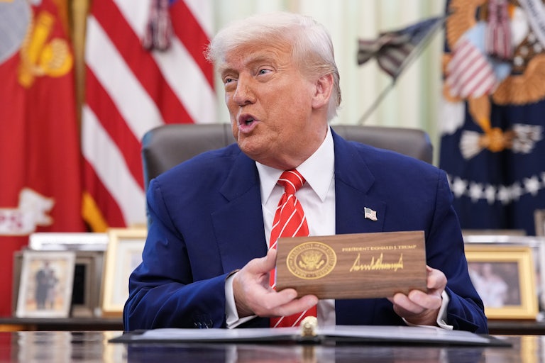 Donald Trump speaks and holds a box with the presidential seal and his signature on it, while sitting at his desk in the Oval Office
