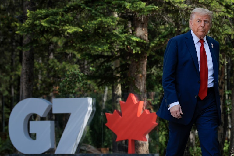 A pissed off looking Trump walks in front of a sign for the G7 summit