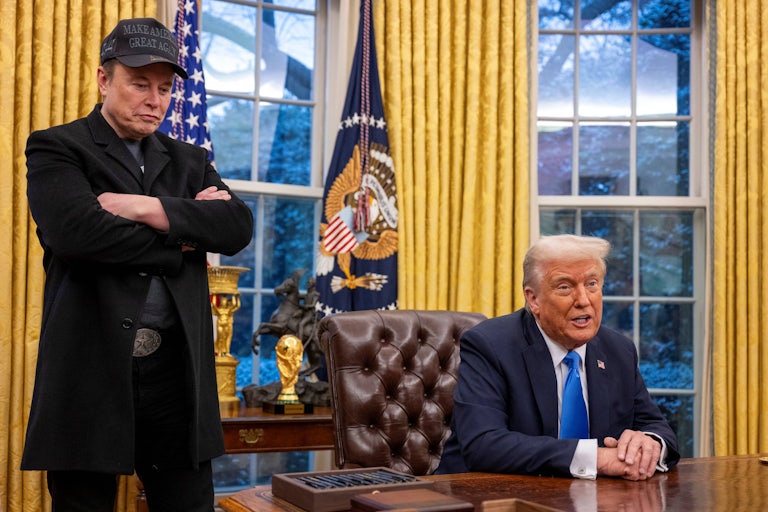 Donald Trump sits at his desk in the Oval Office and speaks during a press conference, as Elon Musk stands next to him with his arms folded