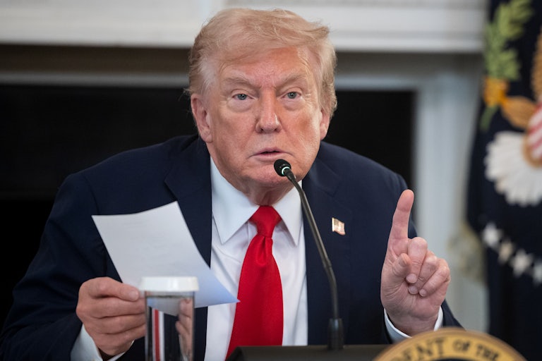 Donald Trump holds a piece of paper in his hand and gestures with his other index finger as he leans in to a mic on the table in front of him.