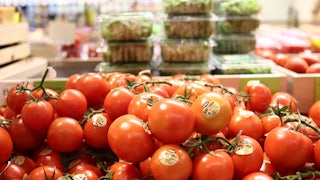 Tomatoes sit in a bin with packaged greens behind them.