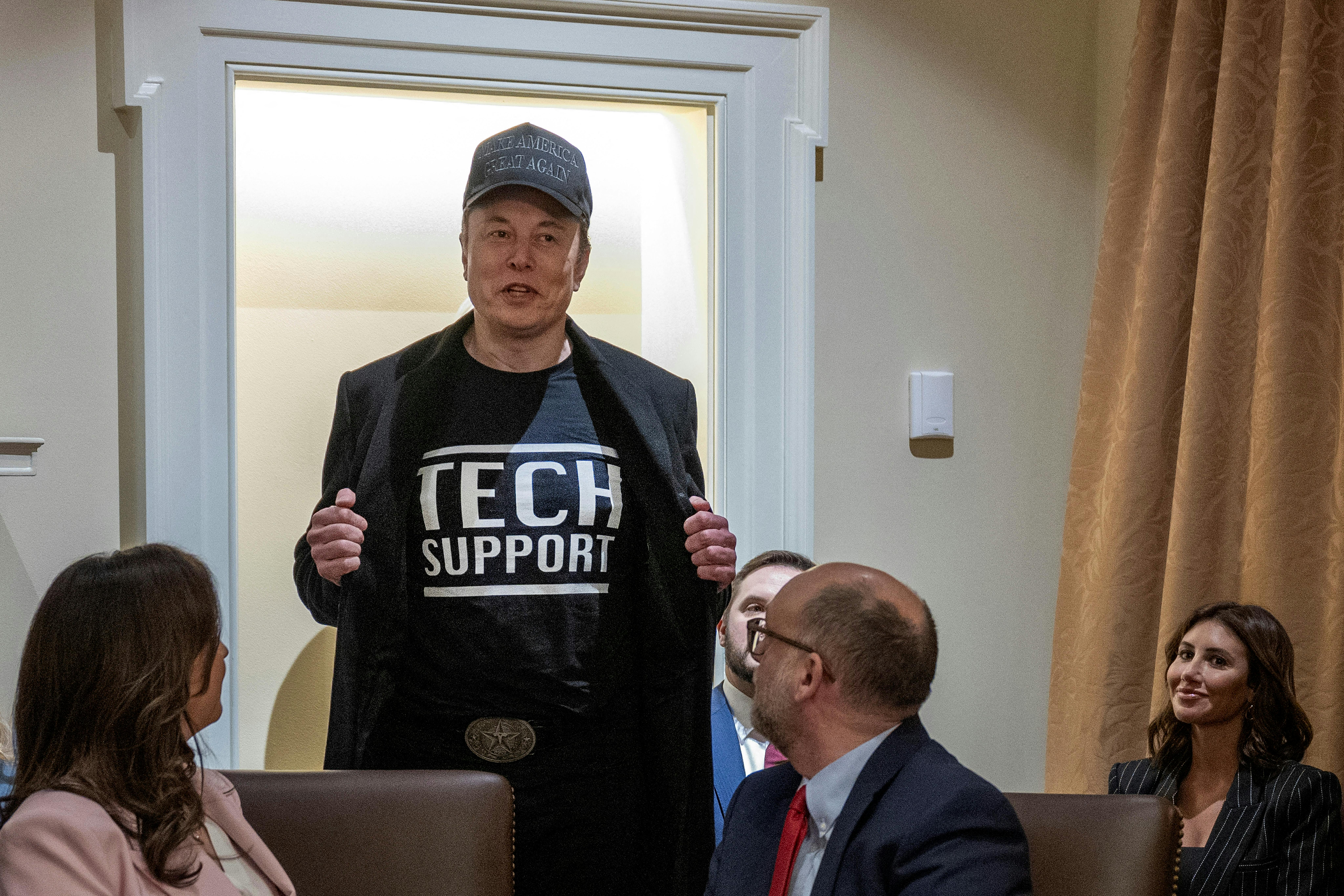 Elon Musk opens his blazer jacket, revealing a shirt that reads "Tech Support." Others sitting around a conference table turn back to look at him.