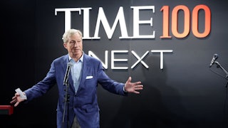 Tom Steyer, wearing a blue blazer, light blue dress shirt, and grey pants, holds his hands out while standing at a microphone and speaking, in front of a background reading "Time100 Next."