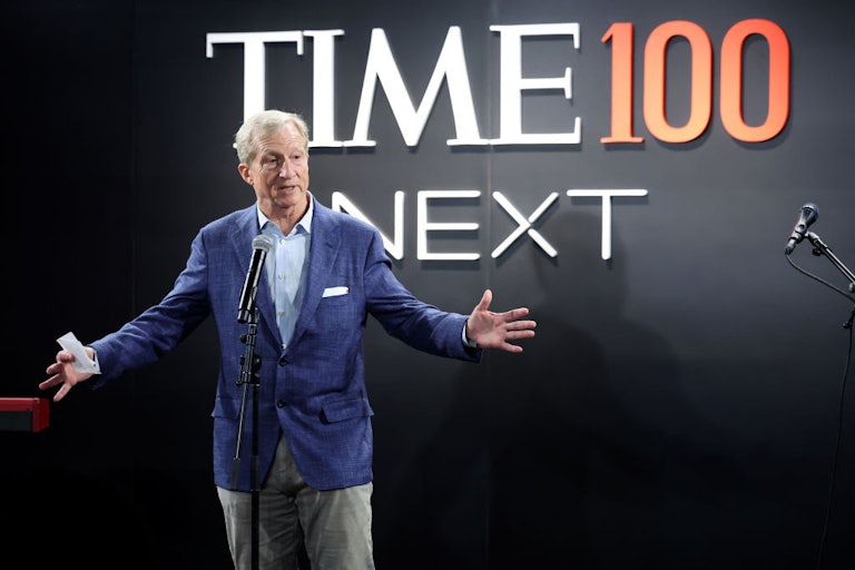 Tom Steyer, wearing a blue blazer, light blue dress shirt, and grey pants, holds his hands out while standing at a microphone and speaking, in front of a background reading "Time100 Next."