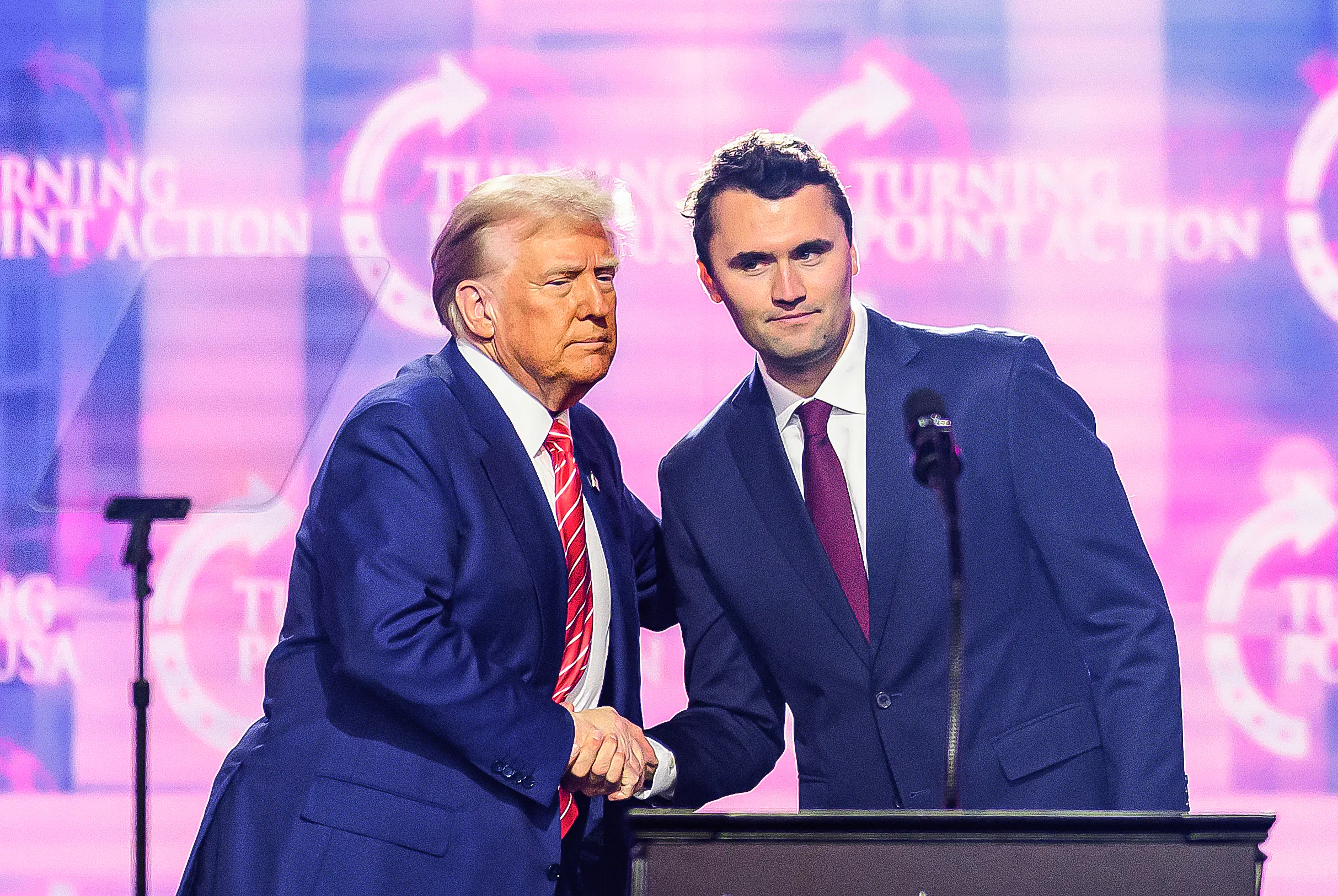 Donald Trump and Charlie Kirk shake hands while standing on stage during a Turning Point USA event