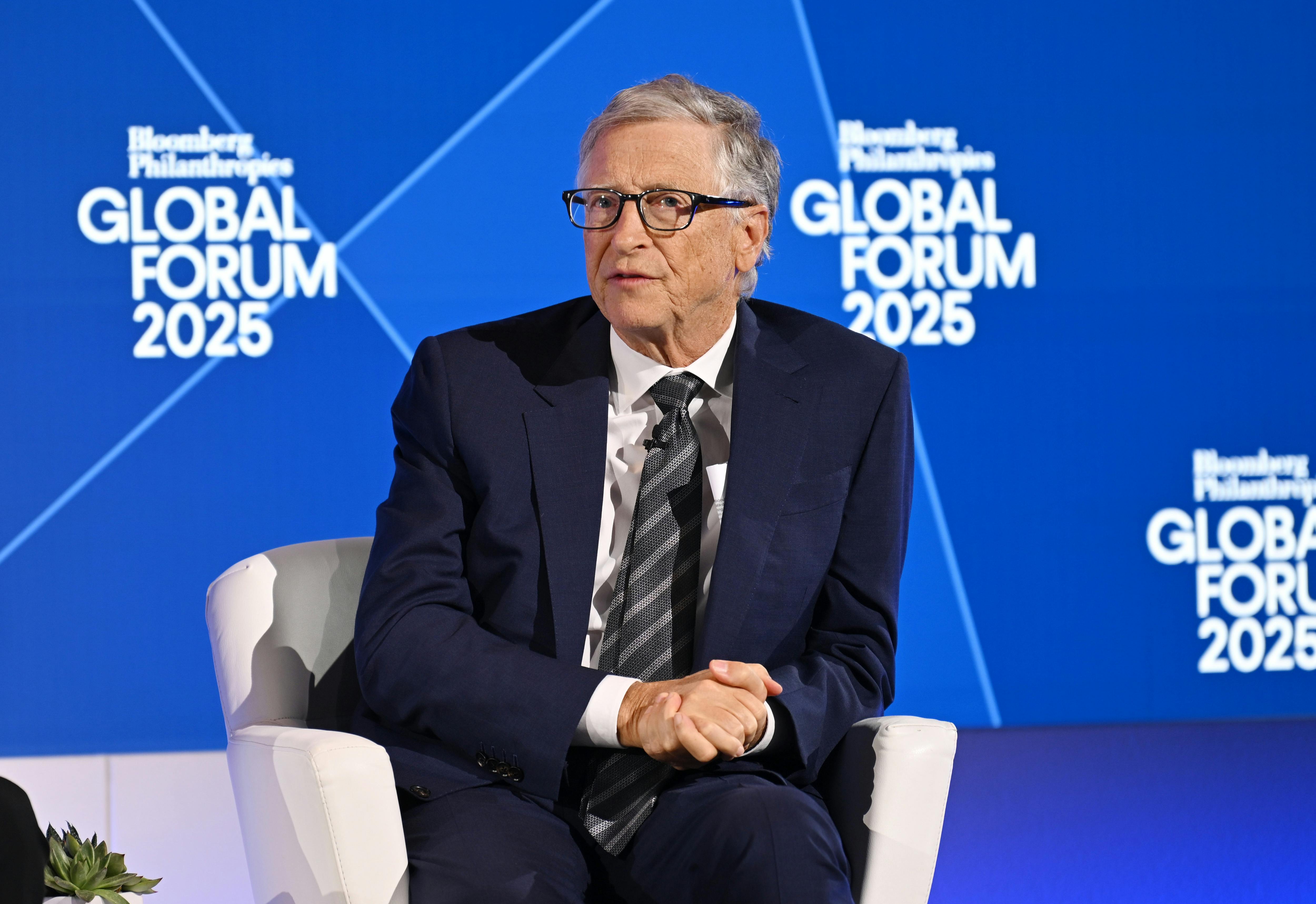 Bill Gates, dressed in a navy suit, sits in a white chair, with his hands clasped, in front of a blue background.
