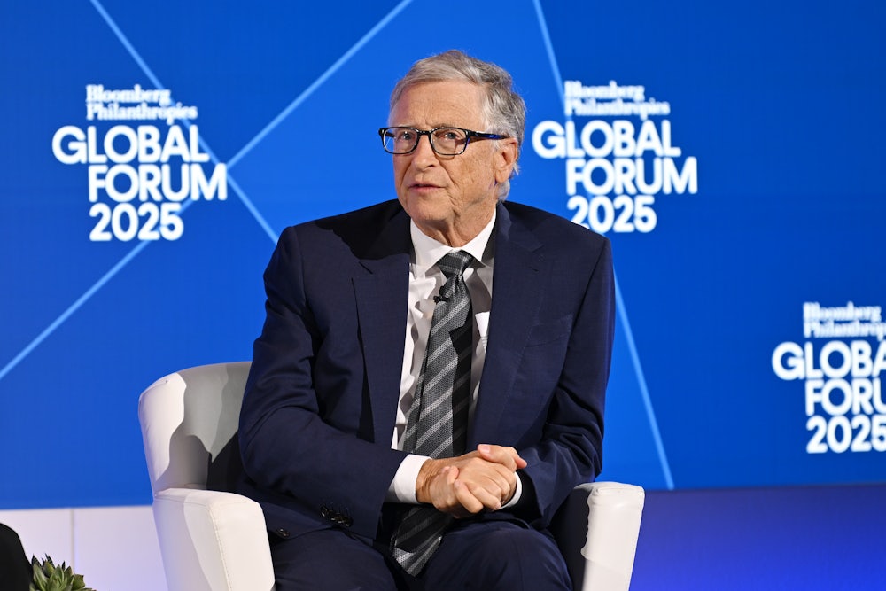 Bill Gates, dressed in a navy suit, sits in a white chair, with his hands clasped, in front of a blue background.