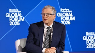 Bill Gates, dressed in a navy suit, sits in a white chair, with his hands clasped, in front of a blue background.