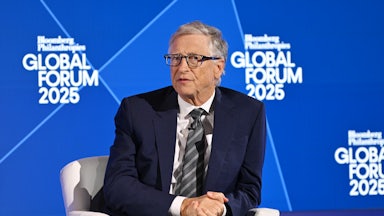 Bill Gates, dressed in a navy suit, sits in a white chair, with his hands clasped, in front of a blue background.