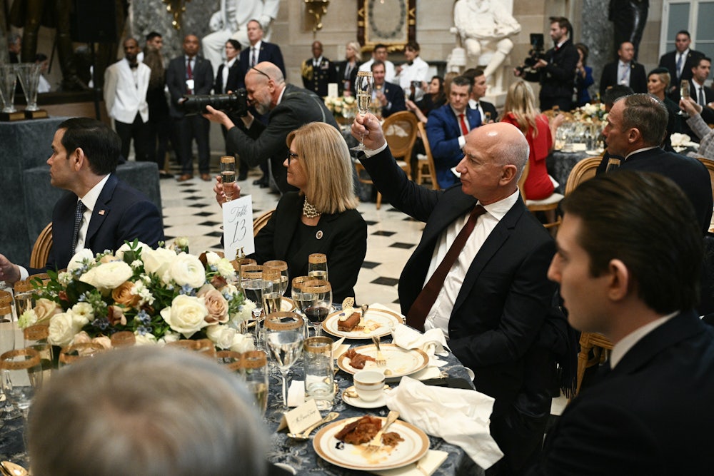 Jeff Bezos sits at a table, raising a champagne flute, while surrounded by others.