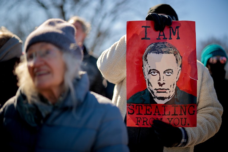 A protester holds a red sign with Elon Musk’s face and the caption "I Am Stealing from You."