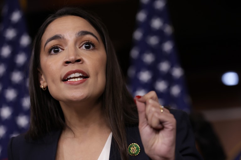 Representative Alexandria Ocasio-Cortez speaking. Two U.S. flags are in the background, out of focus.