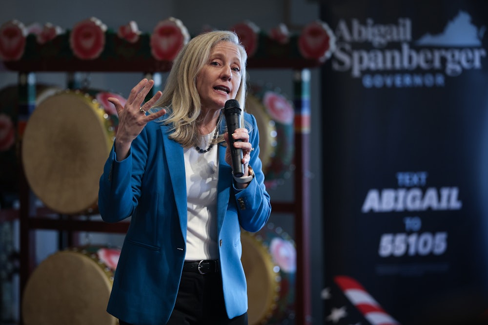 Abigail Spanberger speaks into a microphone while standing in front of a rack with objects on it, while wearing a blue blazer.