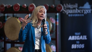 Abigail Spanberger speaks into a microphone while standing in front of a rack with objects on it, while wearing a blue blazer.