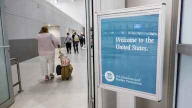 An overweight woman walks past a sign in the airport that says "Welcome to the United States," rolling her carry on luggage.