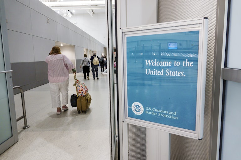 An overweight woman walks past a sign in the airport that says "Welcome to the United States," rolling her carry on luggage.