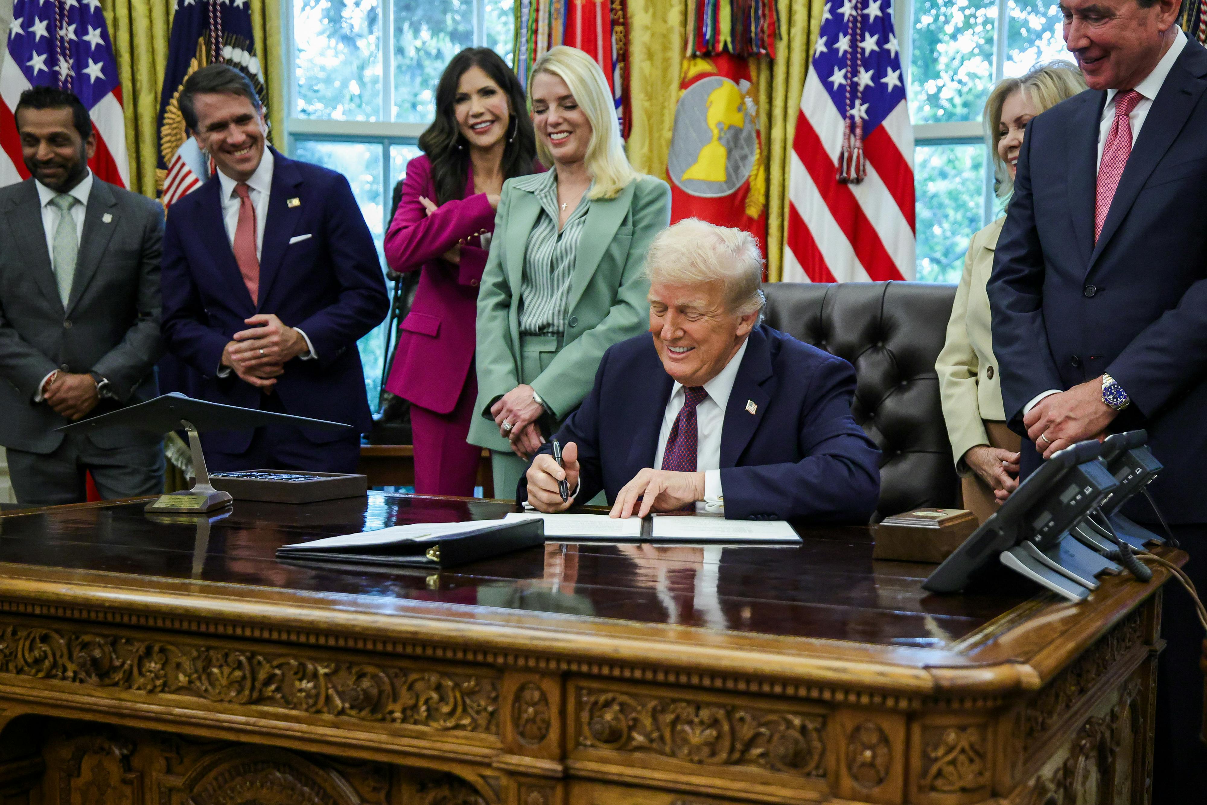 Trump sits at a desk, signing a document, with members of the administration standing behind him.