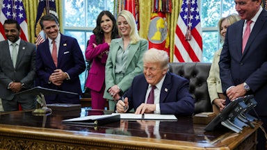 Trump sits at a desk, signing a document, with members of the administration standing behind him.