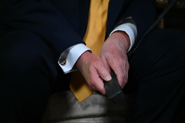 Donald Trump's hands are folded between his knees while he sits. He holds some a phone receiver.