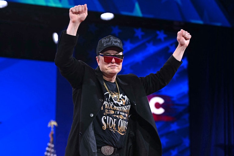 Elon Musk puts both clenched fists in the air as if in victory. He is dressed like a tool with a heavy gold chain, a black MAGA hat, red and black sunglasses, and a graphic tee.