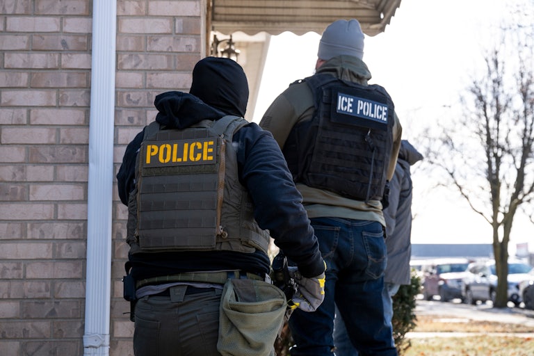 Two people walk towards the front door of a house. They wear sweatshirts, masks, and vests that read "POLICE" and "ICE POLICE" on the back.
