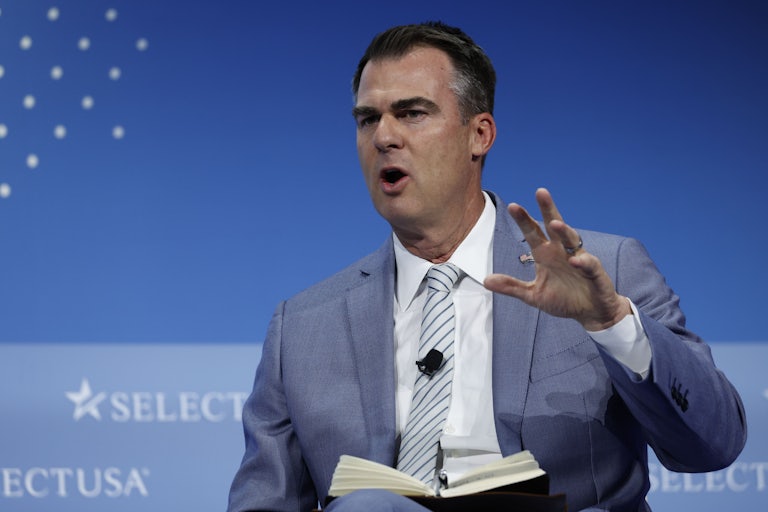 Oklahoma Governor Kevin Stitt wears a suit and speaks and gestures with his hand. A book is on his lap.