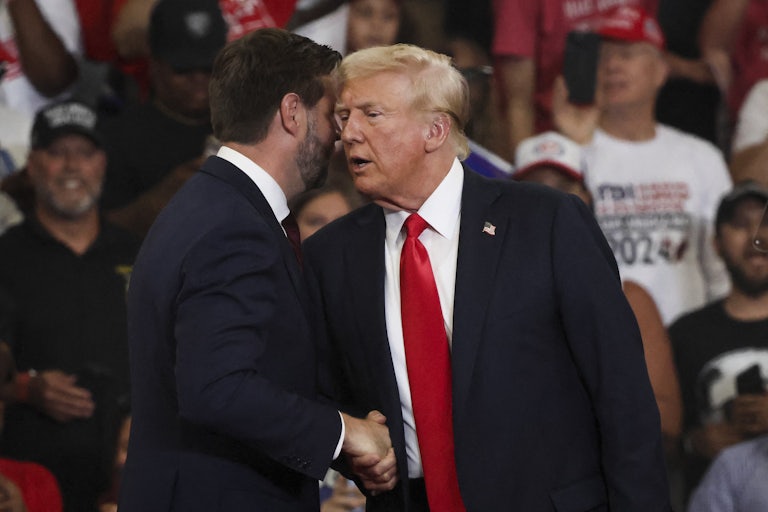Donald Trump shakes J.D. Vance's hand and says something in his ear. Vance's head is turned away from the camera.