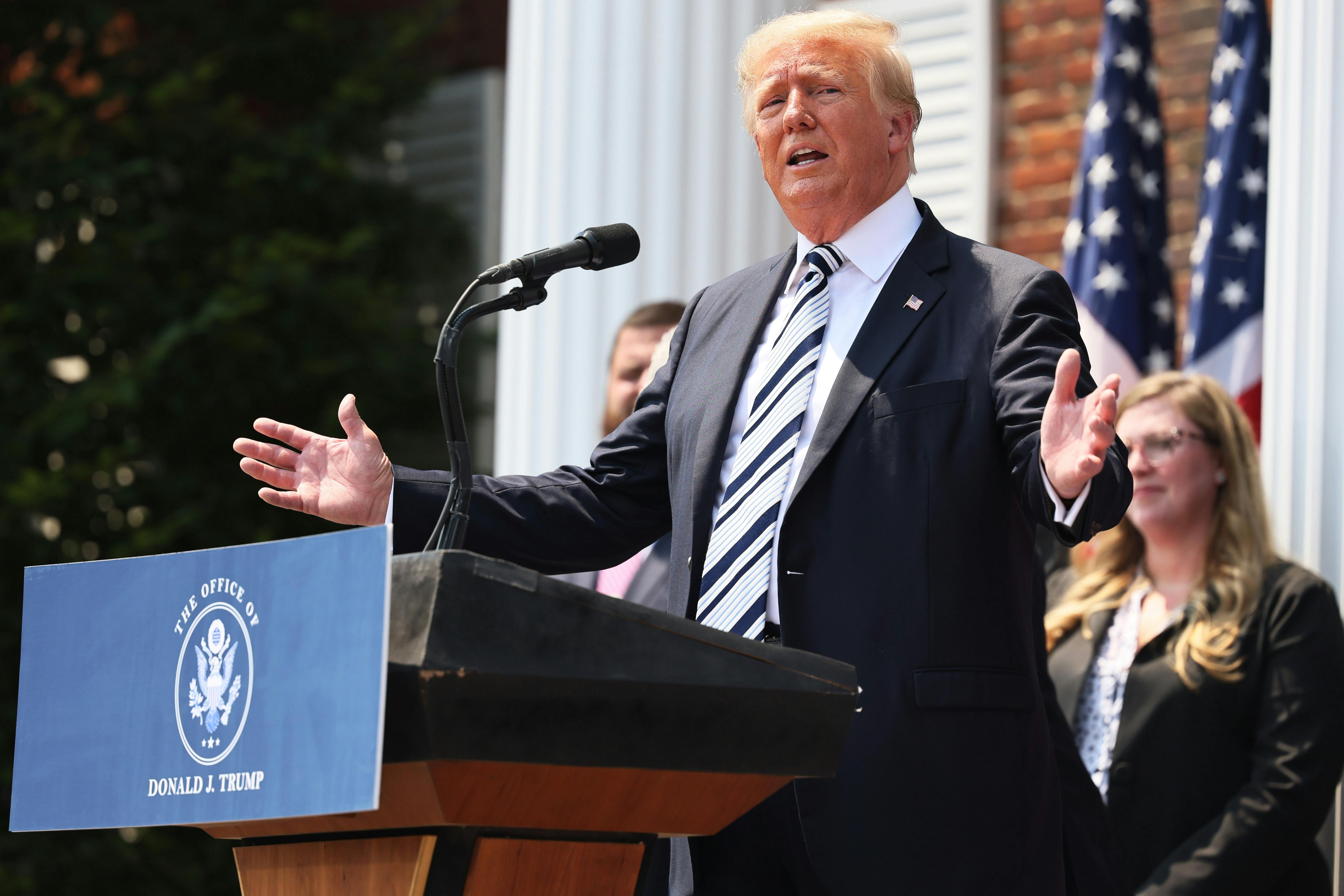 Donald Trump gestures with his hands from behind a podium featuring a sign that reads, "The Office of Donald J. Trump" 