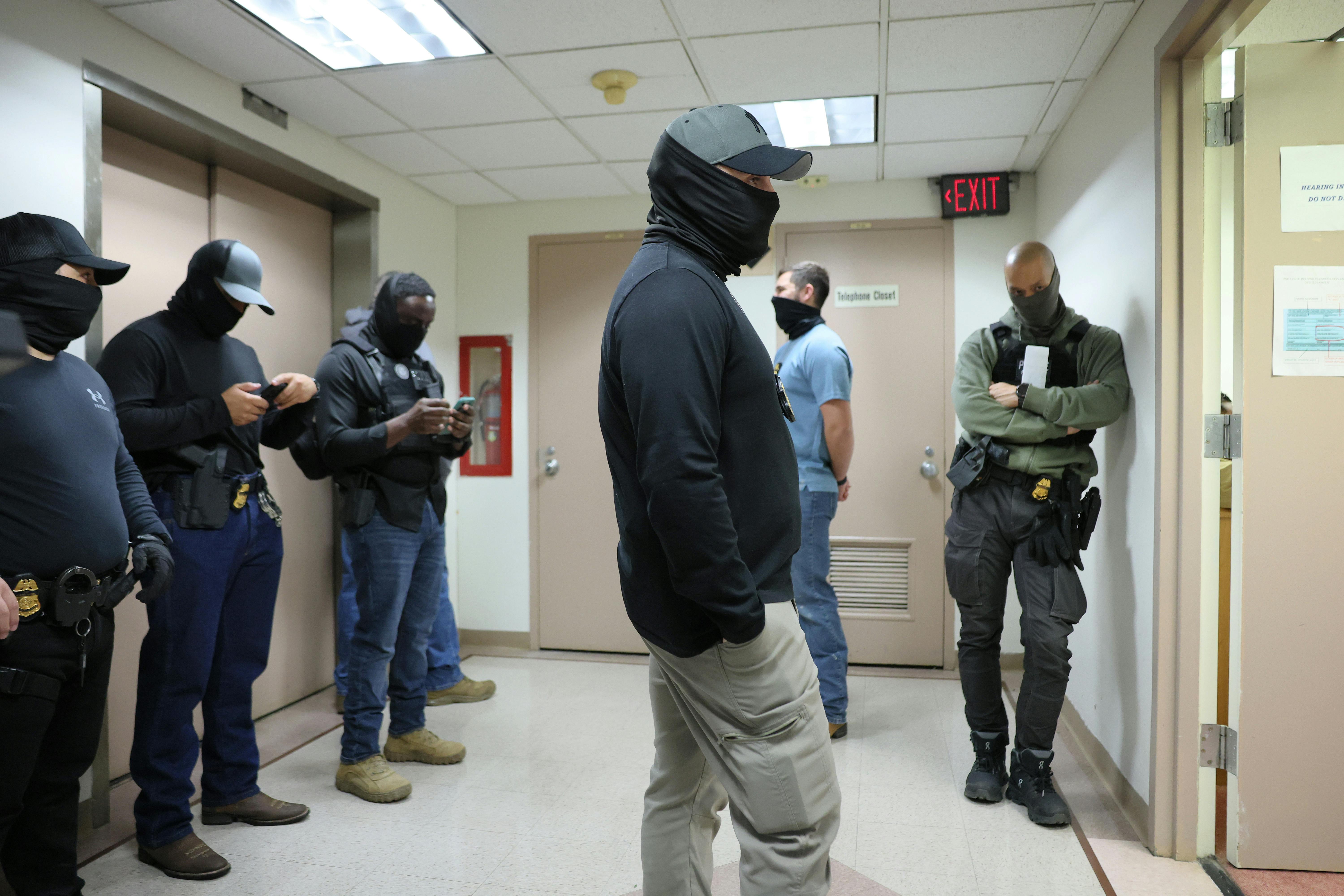Six men stand in a hallway wearing ordinary clothes and balaclavas covering their face.