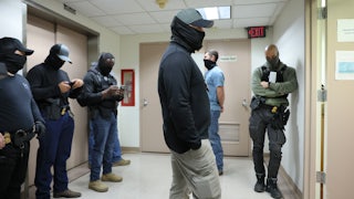 Six men stand in a hallway wearing ordinary clothes and balaclavas covering their face.