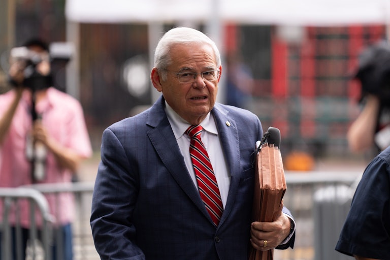 Bob Menendez carries a portfolio in the crook of his arm while walking outside. He appears distressed.