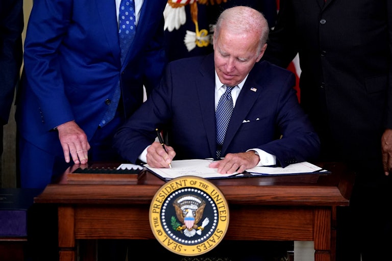 Joe biden signs a bill at a table with the presidential seal.