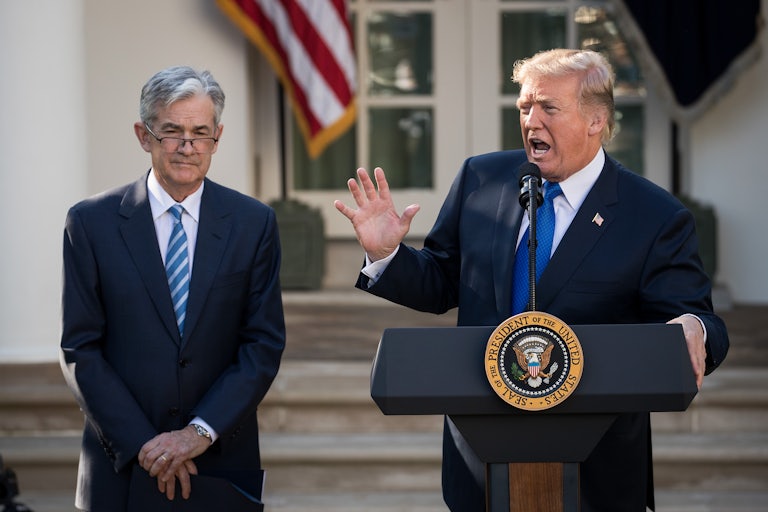 Federal Reserve Chair Jerome Powell stands behind Donald Trump, who gestures and speaks at a podium