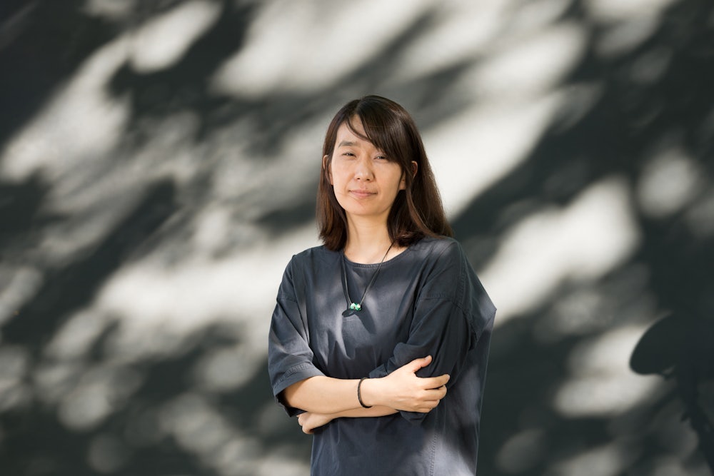 A woman crosses her arms as she stands in front of a shaded wall wearing a black shirt