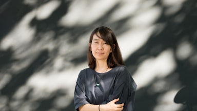 A woman crosses her arms as she stands in front of a shaded wall wearing a black shirt