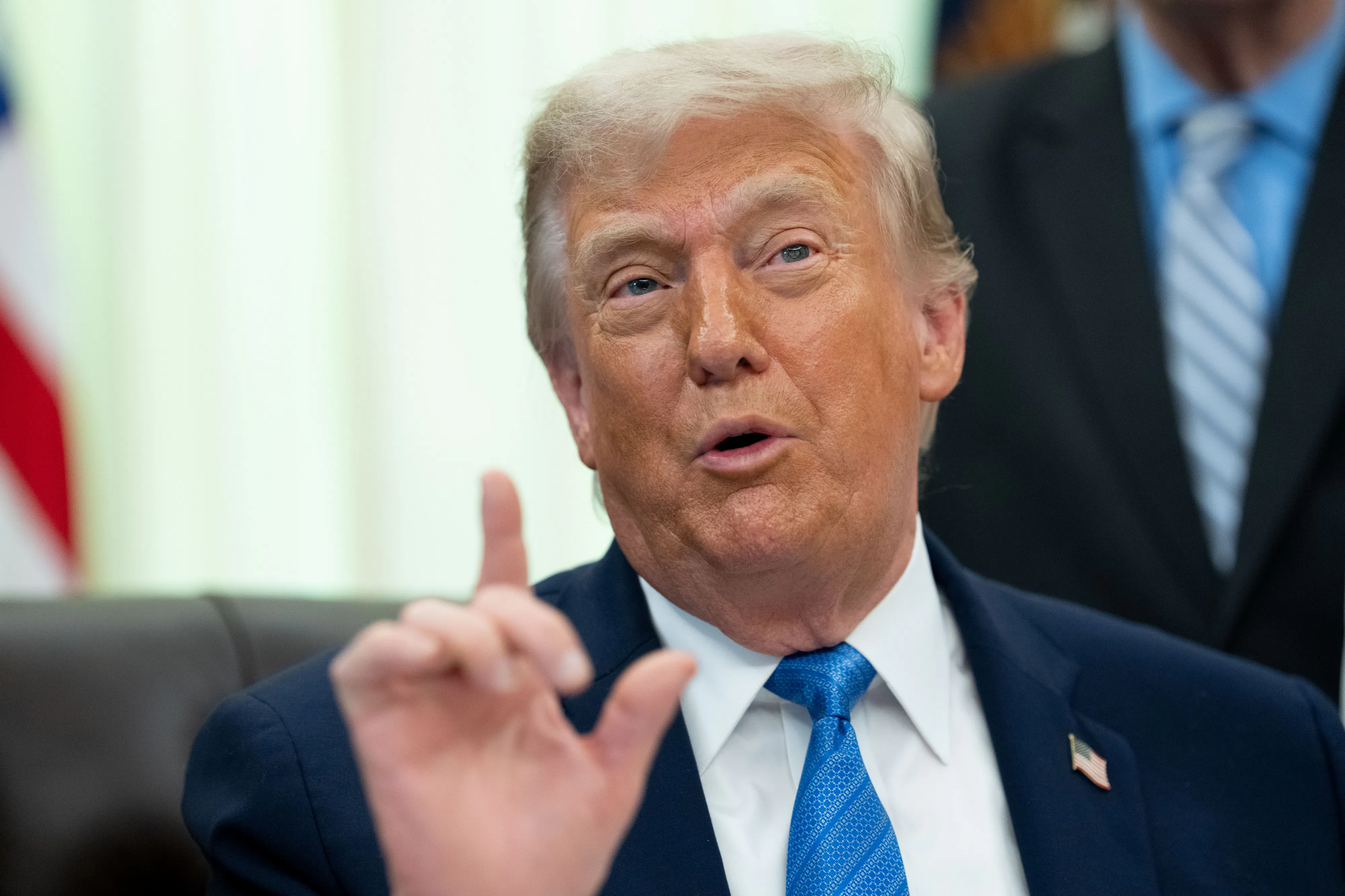 Donald Trump sits at his desk in the Oval Office and speaks, raising one hand with his index finger extended