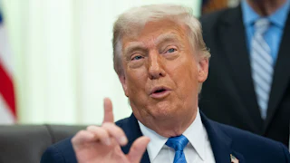 Donald Trump sits at his desk in the Oval Office and speaks, raising one hand with his index finger extended
