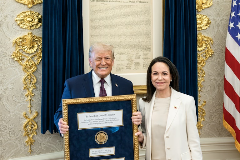 Nobel Peace Prize winner María Corina Machado smiles next to Donald Trump in the Oval Office of the White House, as he holds her framed Nobel Prize.
