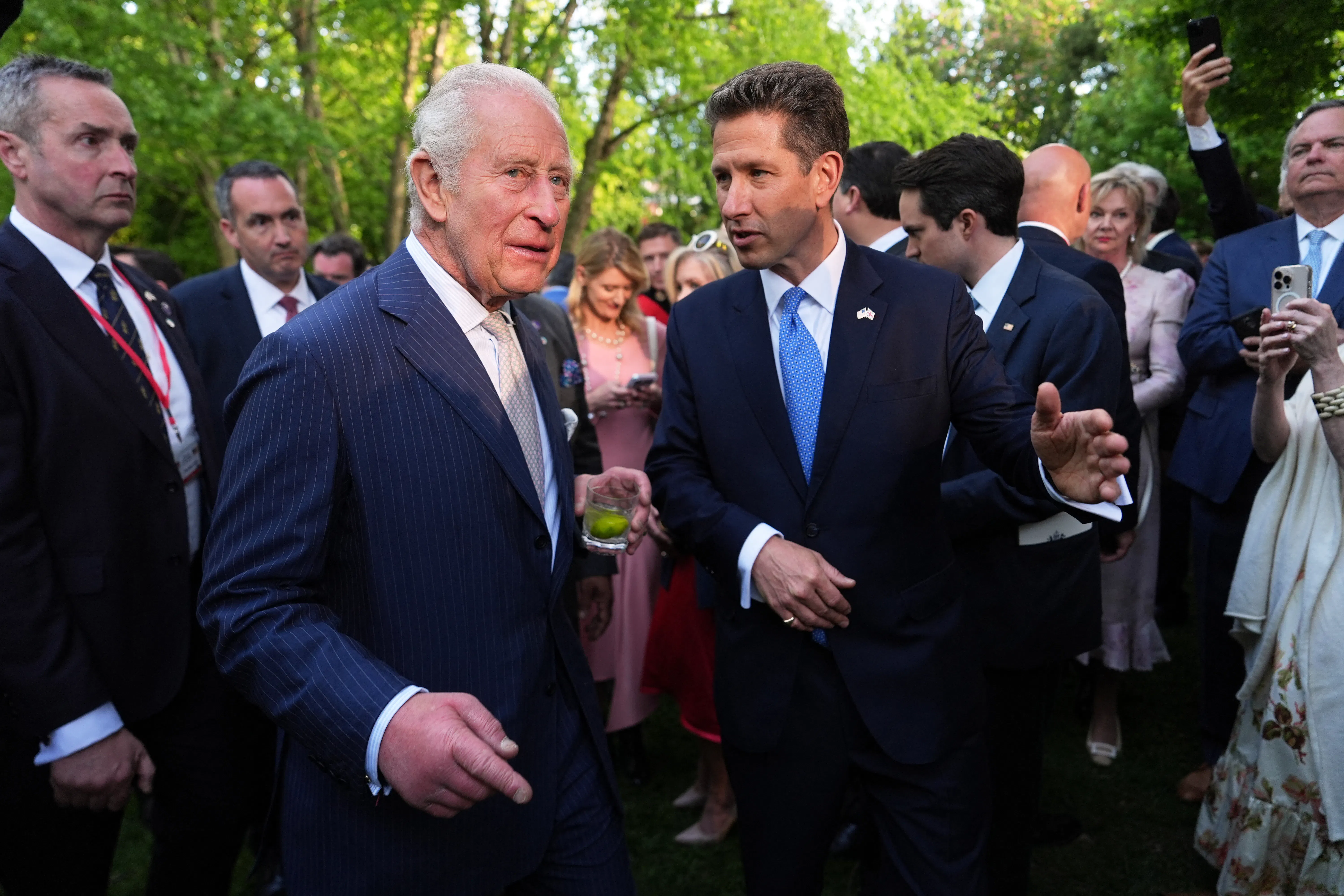 Britain’s King Charles III, drink in hand, and the British Ambassador to the U.S. Christian Turner speak as a group of people surround them.