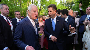 Britain’s King Charles III, drink in hand, and the British Ambassador to the U.S. Christian Turner speak as a group of people surround them.