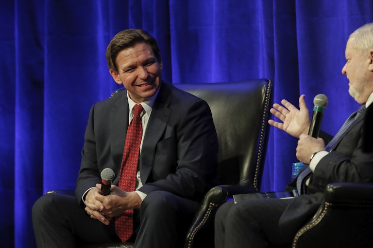 Ron DeSantis smiles while seated and holding a mic, in conversation with someone across from him.