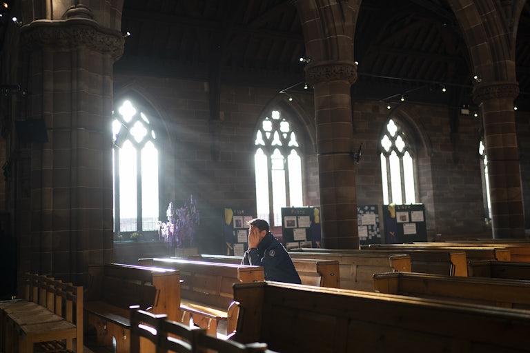 A man sits in the pew of a church praying as sunlight streams through a stained glass window.