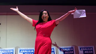 Analilia Mejia raises her arms and smiles with her mouth open while walking on stage. She holds a piece of paper in one hand.
